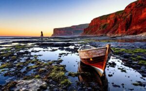 petit-bateau-en-bois-pres-de-hautes-falaises-rouges-au-coucher-du-soleil-eau-calme-avec-reflets-rochers-et-herbes-marines-ciel-orange-et-atmosphere-douce-et-chaleureuse