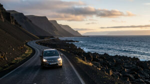 Voiture de location roulant sur une route volcanique noire entre les montagnes et l’océan islandais au lever du jour.