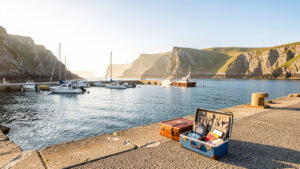 Vue lumineuse de la côte bretonne : petit port, falaises en arrière-plan, valises ouvertes sur le quai et ambiance de week-end marin.