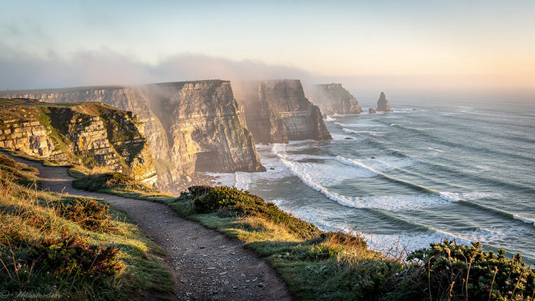 Vue panoramique du littoral breton au matin : falaises et sentier côtier, lumière douce sur la mer et brume légère.